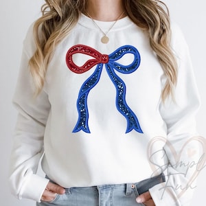 May include: White sweatshirt featuring a large, embroidered bow. The bow has a red top section and blue lower sections, both with glittery accents. The design is centered on the chest, offering a festive and patriotic look.