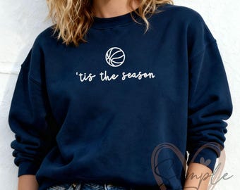 Könnte beinhalten: Marineblaues Sweatshirt mit weißem Basketball-Motiv und dem Schriftzug "'tis the season". Das Sweatshirt hat einen Rundhalsausschnitt und wird mit blauen Jeans getragen.