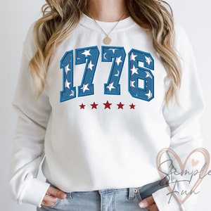 Op de afbeelding: Wit sweatshirt met een blauwe en witte 1776-afbeelding met sterren. De cijfers zijn rood omrand en er staan vijf rode sterren onder de cijfers.