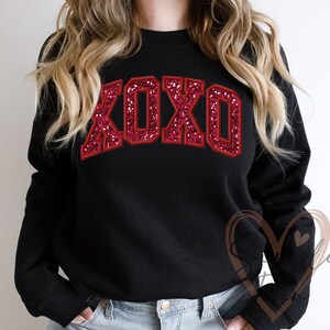 May include: Black sweatshirt with a red glitter XOXO graphic. The sweatshirt has a crew neck and long sleeves.