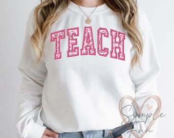 May include: White sweatshirt with the word "TEACH" in pink letters with a floral pattern. The letters are outlined in a darker pink.