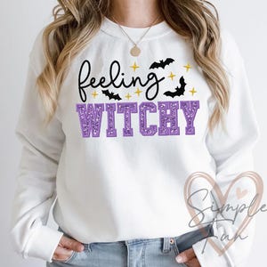 May include: White sweatshirt with the words "feeling witchy" in black script and purple block letters. The design includes black bats and yellow stars, perfect for Halloween or a witchy aesthetic.