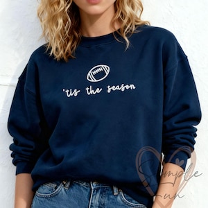 Könnte beinhalten: Marineblaues Sweatshirt mit weißer Football-Grafik und dem Schriftzug "'tis the season". Das Sweatshirt mit Rundhalsausschnitt wird mit einer blauen Jeans kombiniert. Text und Grafik sind auf der Brust zentriert.