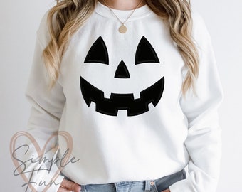May include: White sweatshirt featuring a large, black jack-o'-lantern face. The design includes triangular eyes, a triangular nose, and a wide, toothy grin. The sweatshirt is a crew neck style.