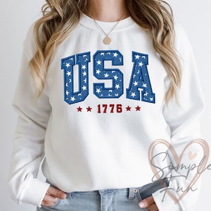 May include: White sweatshirt with a blue and white "USA" graphic with stars. The graphic also includes the year "1776" in red.