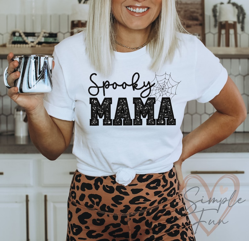 May include: White t-shirt with black glitter text that reads "Spooky Mama" and a black spiderweb graphic.