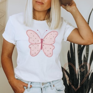 May include: White t-shirt with a large pink butterfly design. The butterfly has a pink outline, pink body, and wings filled with small gold stars. The shirt is a classic crew neck style.