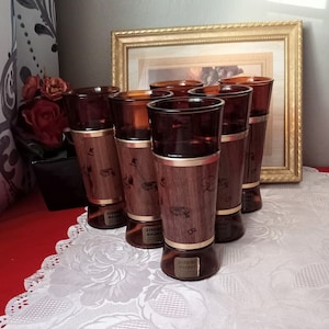 May include: Set of six vintage brown glass tumblers with wooden sleeves and gold trim. The wooden sleeves feature a decorative design. Each glass has a "Genuine Walnut" sticker. The glasses are arranged on a white lace tablecloth.