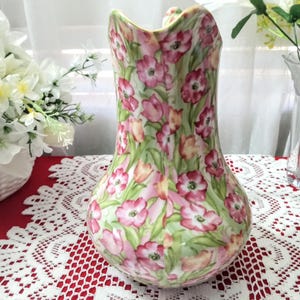 May include: A decorative ceramic pitcher with a floral pattern of pink flowers and green leaves. The pitcher has a curved handle and a wide opening. The background is a red tablecloth with a white lace overlay.