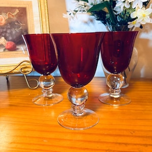 May include: Three ruby red wine glasses with clear, spherical bases and flared rims. The glasses are arranged on a wooden surface, with a framed picture and a vase of white flowers in the background. The glasses are elegant and decorative.