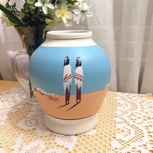 May include: A hand-painted ceramic vase with a light blue sky and tan landscape design. Two figures in traditional attire are depicted. The vase has a white base and rim, and stands approximately 6 inches tall.