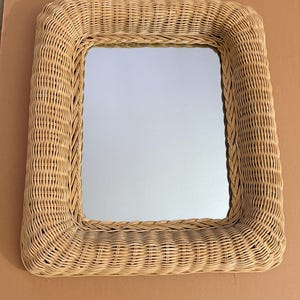 May include: Square woven wicker mirror with a light brown finish. The mirror has a rectangular reflective surface and a thick, rounded frame. The woven texture adds a natural, rustic aesthetic. The mirror is suitable for home decor.