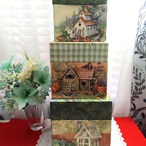 Puede incluir: Una pila de tres cajas decorativas con tapas. Cada caja presenta una escena de jardín diferente con casas para pájaros, flores y pájaros. Las cajas son verdes y tienen una mezcla de patrones, incluido un diseño de cuadros.