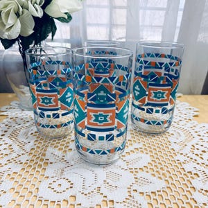 May include: Four clear drinking glasses with a geometric pattern in teal, orange, and navy blue. The glasses are cylindrical and sit on a white lace tablecloth. A vase of white flowers is in the background.