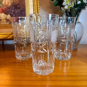 May include: Four clear, cut-glass tumblers with starburst and geometric patterns. The glasses are arranged on a wooden surface, reflecting light. The tumblers are tall and cylindrical, showcasing intricate designs.