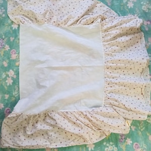 Puede incluir: Una falda de cama blanca con estampado floral rosa y blanco. La falda tiene un borde con volantes y está hecha de una tela suave y ligera.