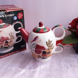 Op de afbeelding: Een witte theepot van keramiek met een rood, groen en wit ontwerp van Sinterklaas en rendieren. De theepot is onderdeel van een thee voor één set. De doos zegt "Santa's Spirit" en "Painted Tea For One".