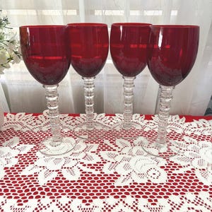 Puede incluir: Cuatro copas de vino tinto con tallos y bases transparentes y texturizados. Los cuencos rojos tienen rayas horizontales. Las copas están dispuestas sobre una superficie roja cubierta con un mantel de encaje blanco.