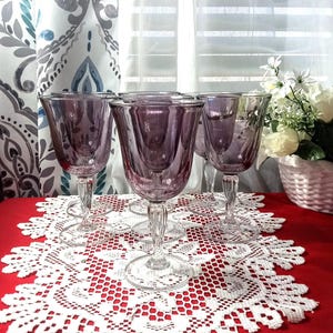 May include: Six purple-tinted wine glasses with clear, twisted stems are displayed on a white lace doily. The glasses sit on a red surface. The background features a patterned curtain and a floral arrangement.