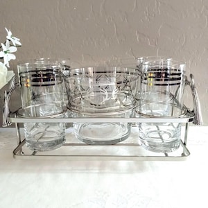 May include: A vintage silver-toned serving set with six clear glass cups and a central bowl. The cups have silver bands and the bowl has a decorative etched design. The set is on a silver metal tray with handles, perfect for serving drinks.