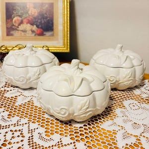 May include: Three off-white ceramic lidded containers shaped like pumpkins. Each container features a ribbed surface with embossed leaf and vine scrollwork details around the base and a sculpted stem handle on the lid, finished in a glossy glaze.