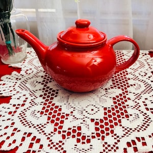 Może przedstawiać: Błyszczący, czerwony ceramiczny imbryk z pasującą pokrywką i uchwytem. Imbryk stoi na białej koronkowej serwetce na czerwonej powierzchni. W tle widoczny jest szklany wazon z kwiatami.