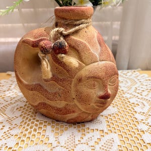 Puede incluir: Un jarrón de cerámica de color terracota con un diseño de cara de sol. El jarrón está adornado con un detalle de cuerda alrededor del cuello y cuentas decorativas. Flores blancas con centros amarillos están dispuestas dentro.