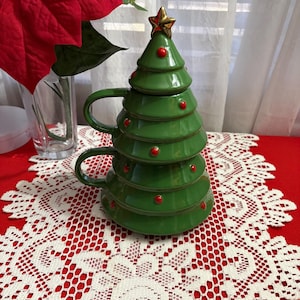 May include: A stackable ceramic mug set shaped like a green Christmas tree with a gold star topper. The design features three tiers, each with a handle and small red circular ornaments. The mugs are glazed in a glossy finish and fit together.