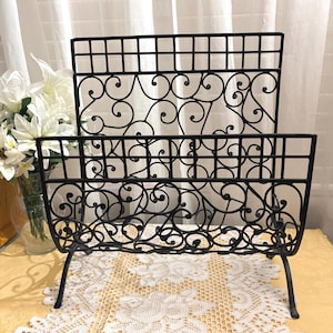 May include: Black metal magazine rack with a decorative scroll design. The rack has a rectangular shape with a grid-like top and a lower section with a curved, openwork pattern. It stands on four curved legs.