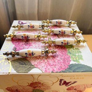 May include: Five drawer pulls with white ceramic centers featuring colorful floral patterns and gold-toned metal ends. The pulls are arranged on a rectangular box decorated with pink hydrangea flowers, green leaves, and a butterfly illustration.