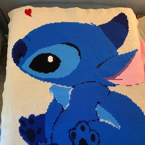 May include: A blue and white crocheted blanket featuring Stitch from Lilo & Stitch. Stitch is sitting with his back to the viewer and has a pink ear. A red heart is in the top left corner of the blanket.
