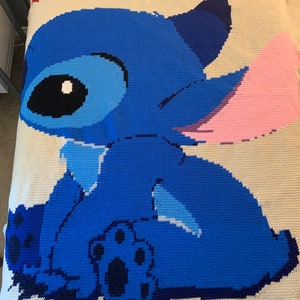 May include: A crocheted blanket featuring a blue Stitch character from the Disney movie Lilo & Stitch. The character is sitting with its back to the viewer and has a pink ear.