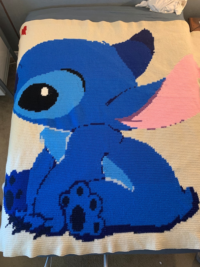 May include: A crocheted blanket featuring a blue Stitch character from the Disney movie Lilo & Stitch. The character is sitting with its back to the viewer and has a pink ear.