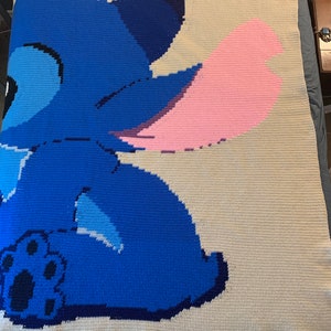 May include: A crocheted blanket featuring a blue Stitch character from the Disney movie Lilo & Stitch. Stitch is sitting with his back to the viewer and his pink ear is visible. The blanket is made with a white background and blue, pink, and black yarn.