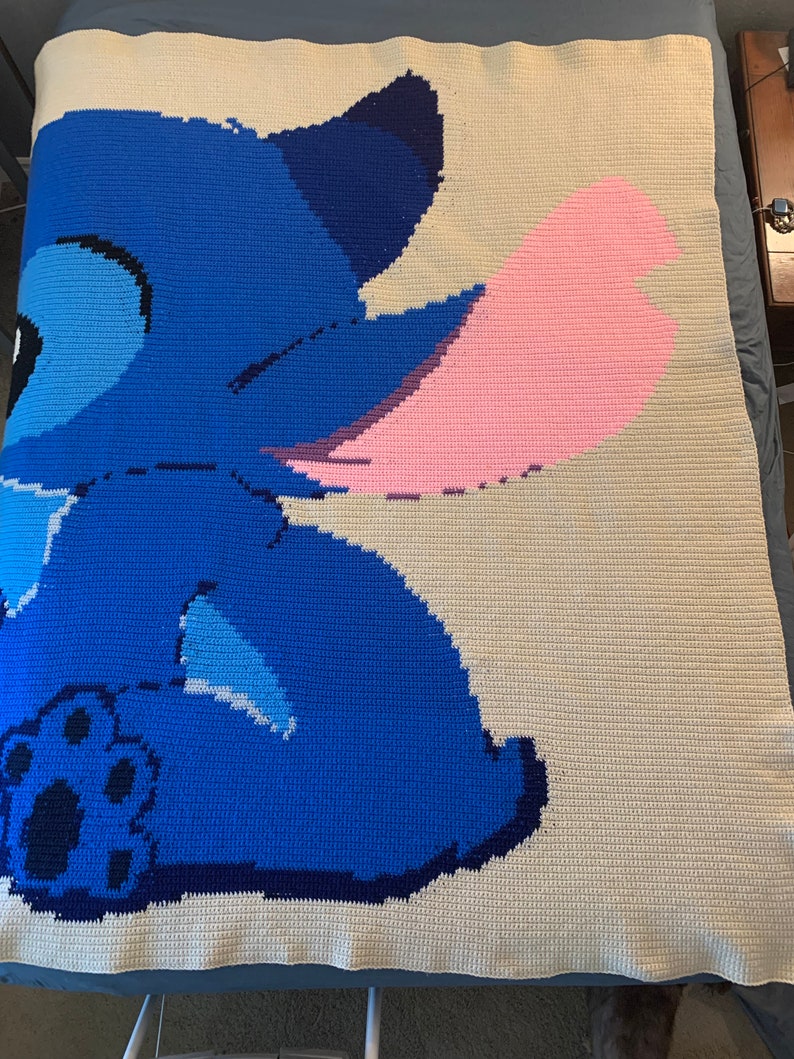 May include: A crocheted blanket featuring a blue Stitch character from the Disney movie Lilo & Stitch. Stitch is sitting with his back to the viewer and his pink ear is visible. The blanket is made with a white background and blue, pink, and black yarn.