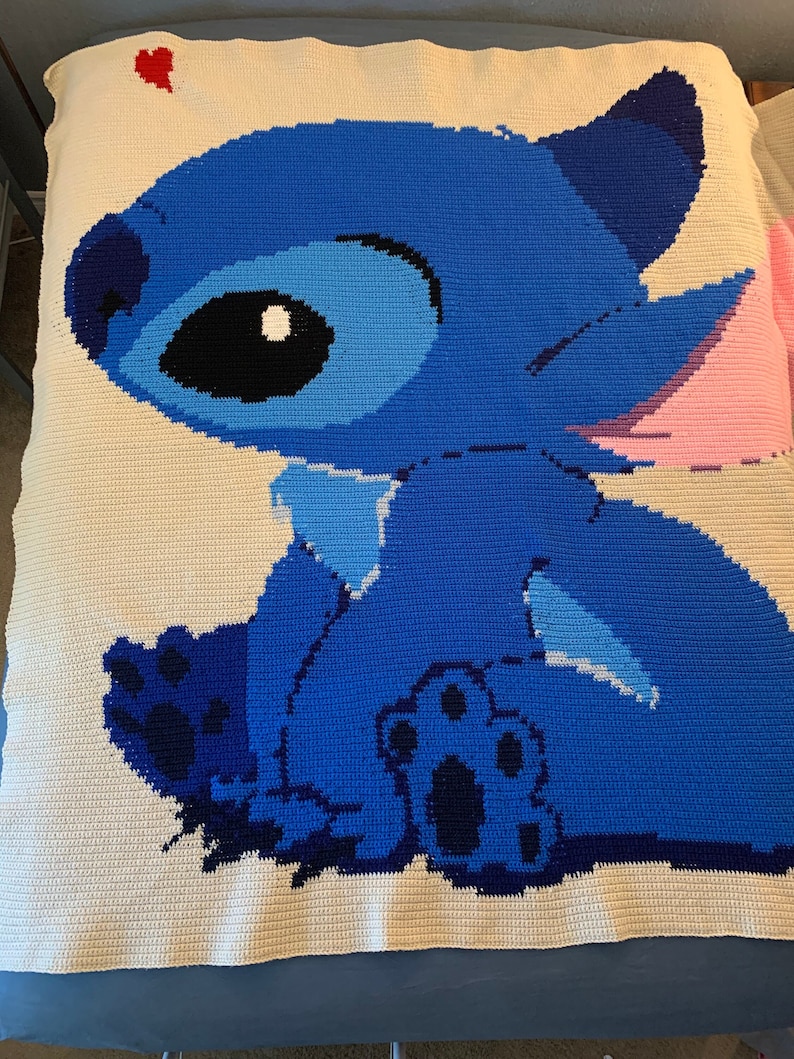 May include: A blue and white crocheted blanket featuring Stitch from Lilo & Stitch. Stitch is sitting with his back to the viewer and has a pink ear. A red heart is in the top left corner of the blanket.