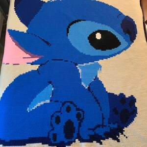May include: A blue and white crocheted blanket featuring Stitch from Lilo & Stitch. Stitch is sitting with his back to the viewer and has a pink ear and a blue paw visible. A red heart is in the upper right corner of the blanket.