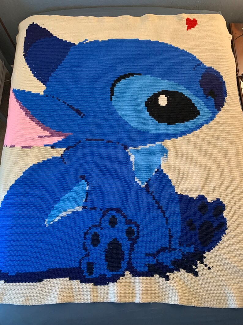May include: A blue and white crocheted blanket featuring Stitch from Lilo & Stitch. Stitch is sitting with his back to the viewer and has a pink ear and a blue paw visible. A red heart is in the upper right corner of the blanket.