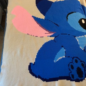 May include: A crocheted blanket featuring Stitch from Lilo & Stitch. Stitch is blue with a pink ear and a black paw print. The blanket is white with a blue and pink design.