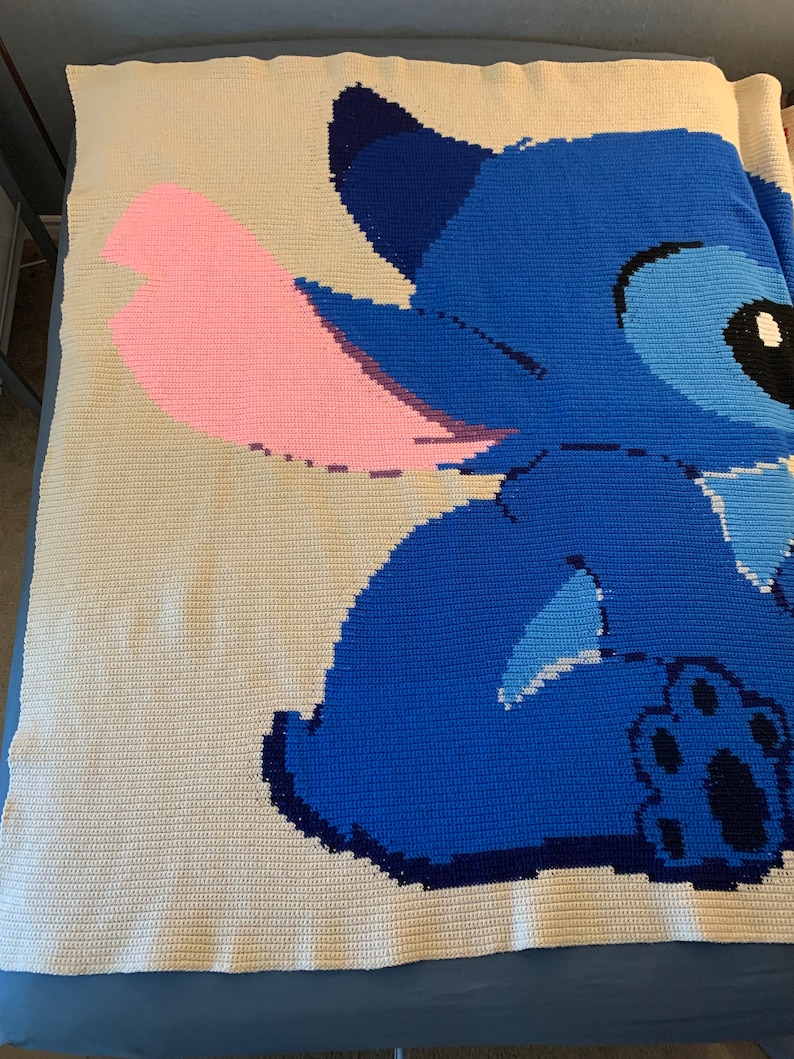 May include: A crocheted blanket featuring Stitch from Lilo & Stitch. Stitch is blue with a pink ear and a black paw print. The blanket is white with a blue and pink design.
