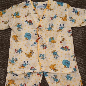 May include: A vintage pajama set featuring a repeating pattern of cartoon characters, including Mickey Mouse and Donald Duck, on a cream-colored background. The set includes a short-sleeved top and pants, trimmed with yellow piping.
