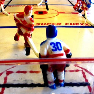 May include: A tabletop hockey game with red, white, and blue players. The game is called "Super Chexx" and features a blue player with the number 30 on their jersey.