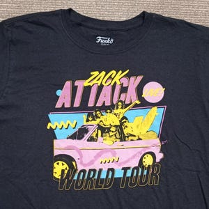 May include: Dark navy blue t-shirt featuring a graphic of a pink car with people inside, the words "Zack Attack World Tour" in yellow and pink, and the Funko logo. The shirt has short sleeves and a crew neck.