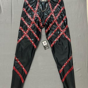 May include: Black trousers with red crisscross detailing and silver trim. The trousers have a silver buckle with a chain and are decorated with small silver studs. A tag is attached near the bottom.