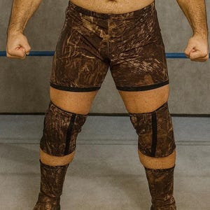 May include: Brown camouflage wrestling shorts and knee pads with black trim. The shorts are above the knee, and the knee pads extend to the mid-calf. The fabric has a mottled pattern of brown and tan.