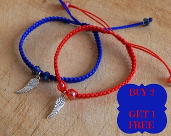 May include: Two braided bracelets with silver wing charms. One bracelet is red and the other is blue. The bracelets are on a wooden surface. The text "BUY 2 GET 1 FREE" is in a blue box.
