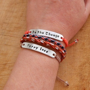 May include: Two bracelets with silver metal tags that say "Be the Change" and "Worry Less". The bracelets are made of braided cords in shades of orange, brown, white, and grey.