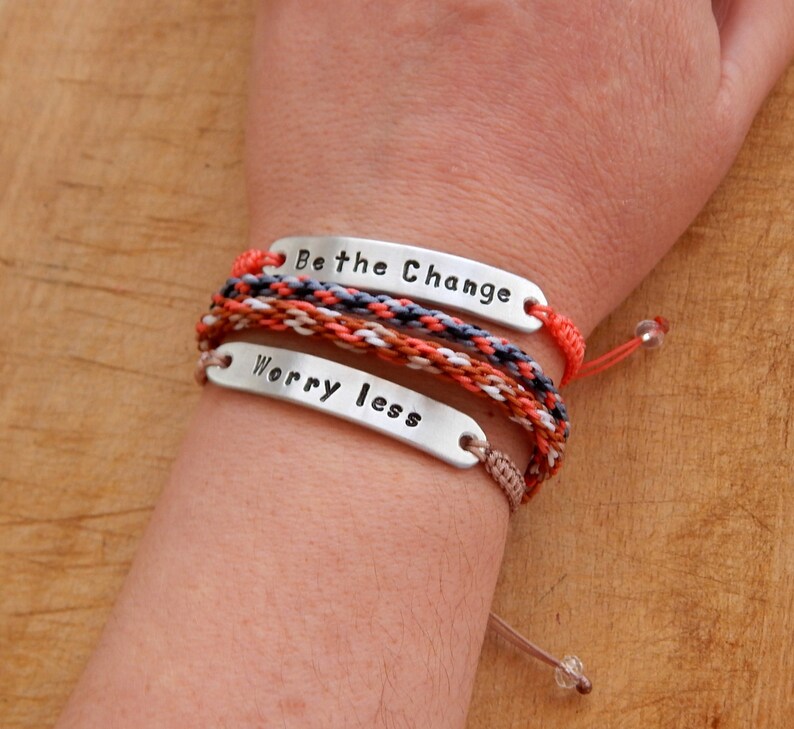 May include: Two bracelets with silver metal tags that say "Be the Change" and "Worry Less". The bracelets are made of braided cords in shades of orange, brown, white, and grey.
