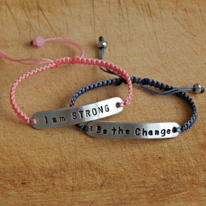 May include: Two adjustable bracelets with silver metal tags. The pink bracelet has the text "I am STRONG" on the tag. The grey bracelet has the text "Be the Change" on the tag.