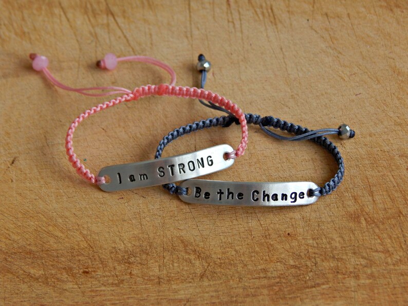 May include: Two adjustable bracelets with silver metal tags. The pink bracelet has the text "I am STRONG" on the tag. The grey bracelet has the text "Be the Change" on the tag.